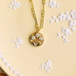 A gold chain necklace with a circular pendant featuring a floral design and three clear stones, displayed on a beige plate with white floral embossing.