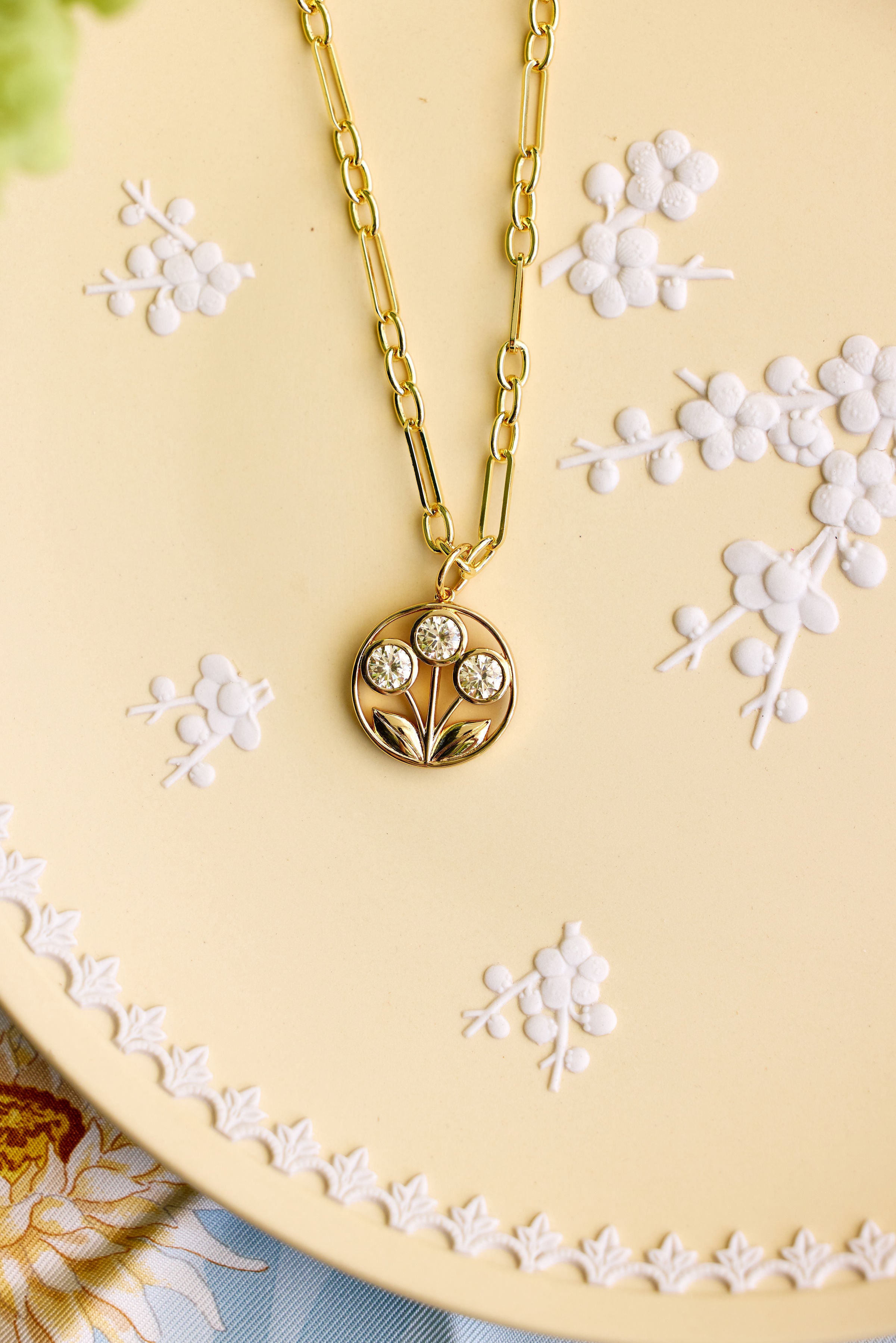 A gold chain necklace with a circular pendant featuring a floral design and three clear stones, displayed on a beige plate with white floral embossing.