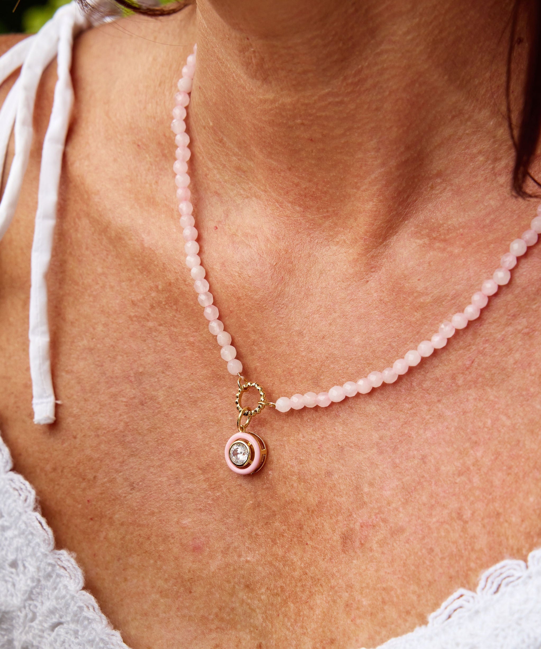 A close-up of a woman wearing a delicate pink beaded necklace with a gold-toned pendant featuring a small white stone, paired with a white lace top.