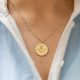 scalloped edge round gold pendant with a floral blossom and green and blue gemstones on a gold chain on a woman's neck