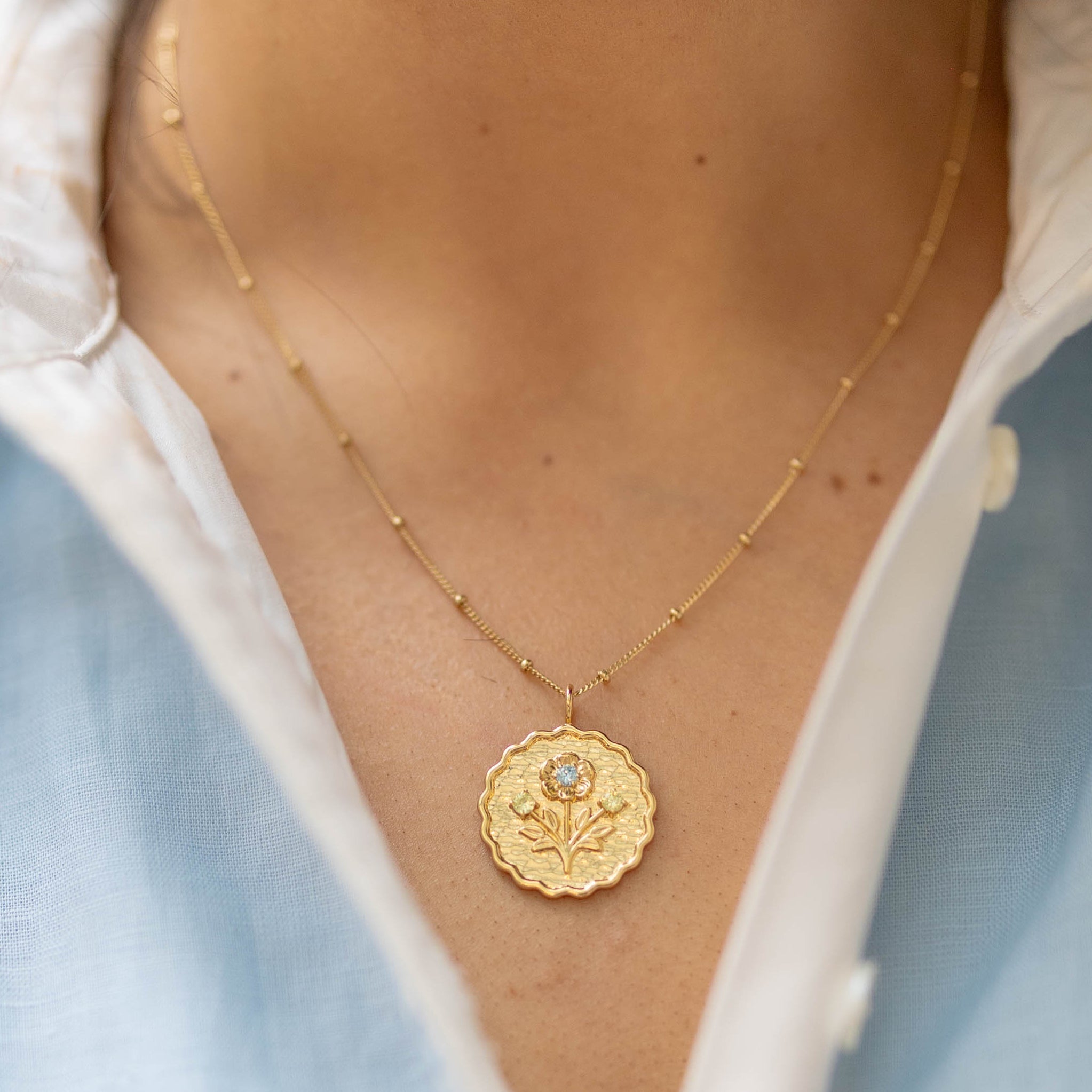 scalloped edge round gold pendant with a floral blossom and green and blue gemstones on a gold chain on a woman's neck