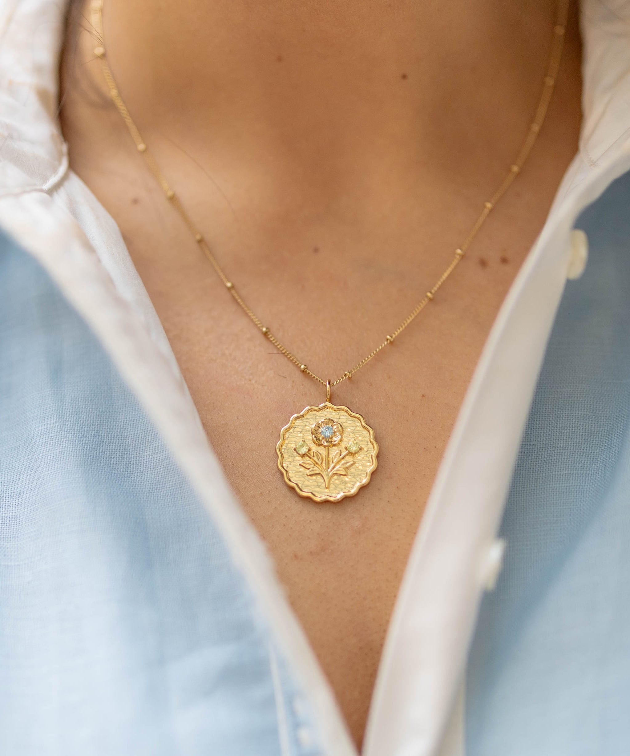scalloped edge round gold pendant with a floral blossom and green and blue gemstones on a gold chain on a woman's neck