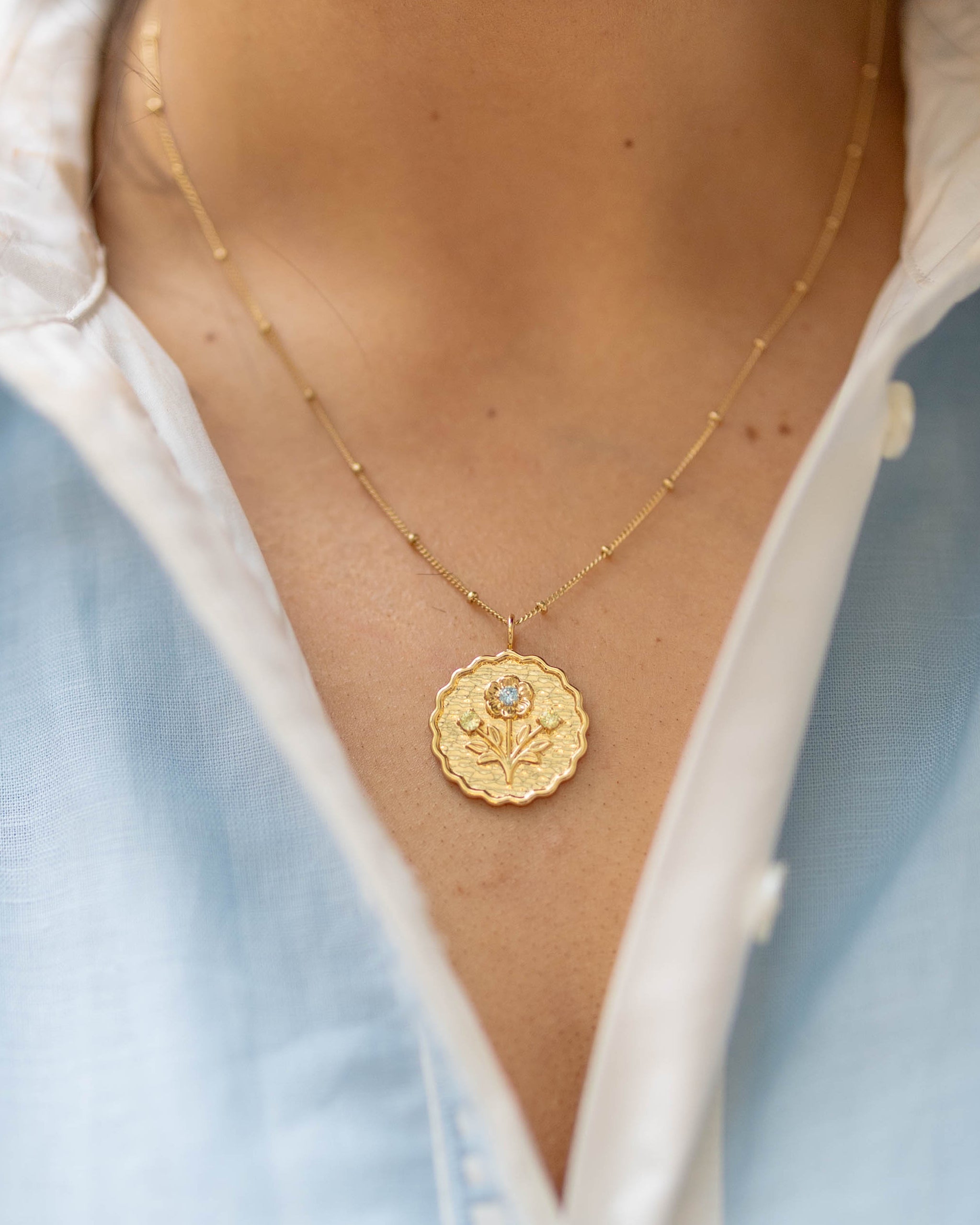 scalloped edge round gold pendant with a floral blossom and green and blue gemstones on a gold chain on a woman's neck