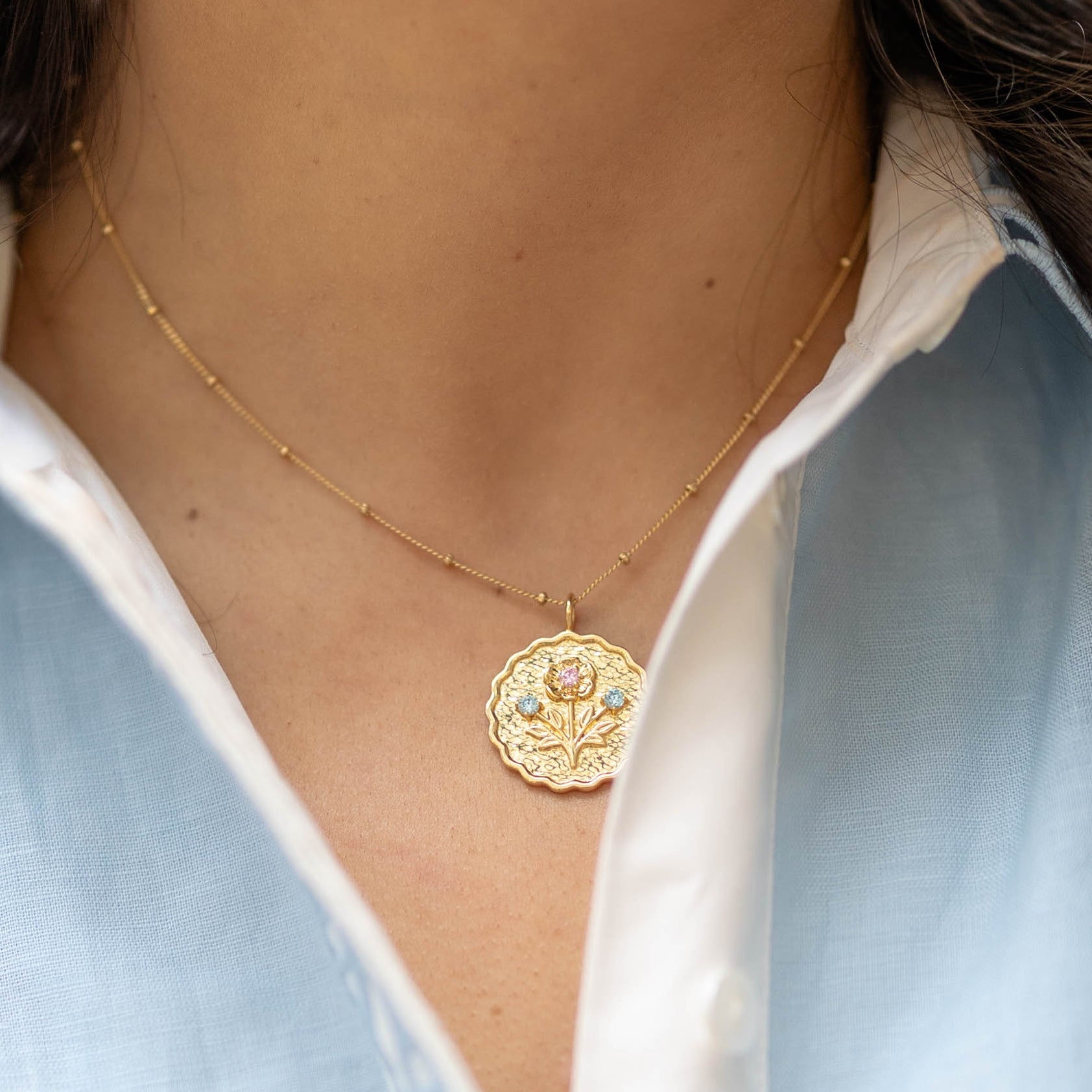 scalloped edge round pendant gold charm with a floral bloom and blue and pink gemstones on a gold chain on a woman's neck