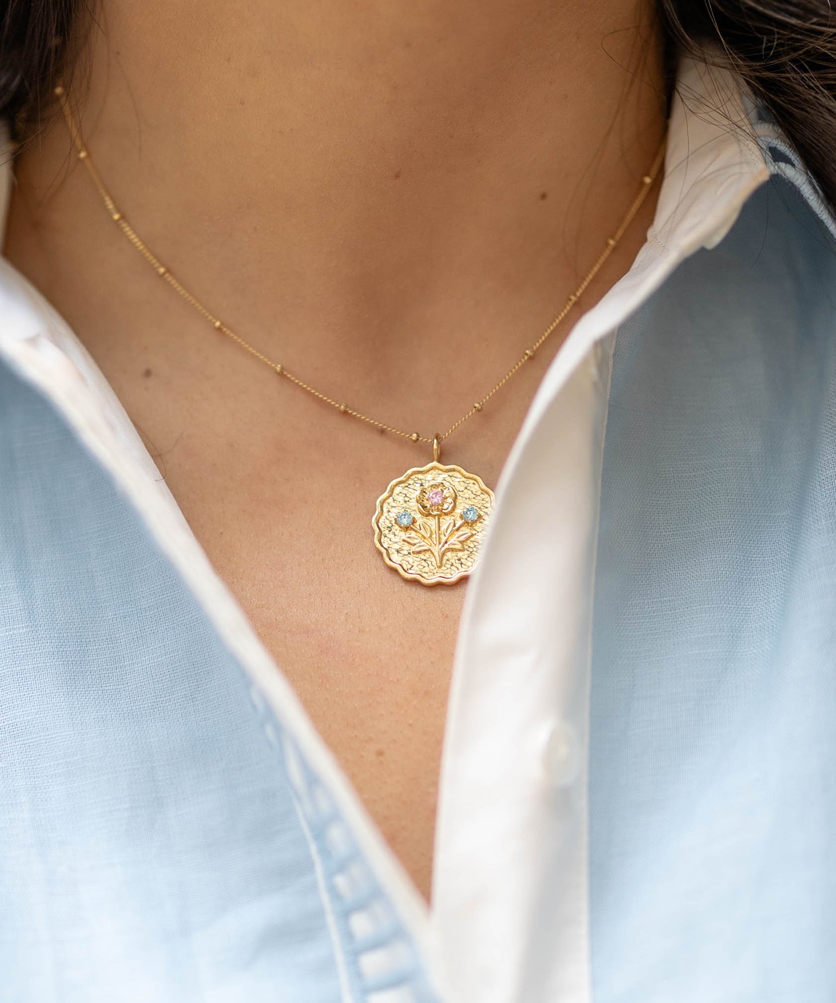 scalloped edge round pendant gold charm with a floral bloom and blue and pink gemstones on a gold chain on a woman's neck