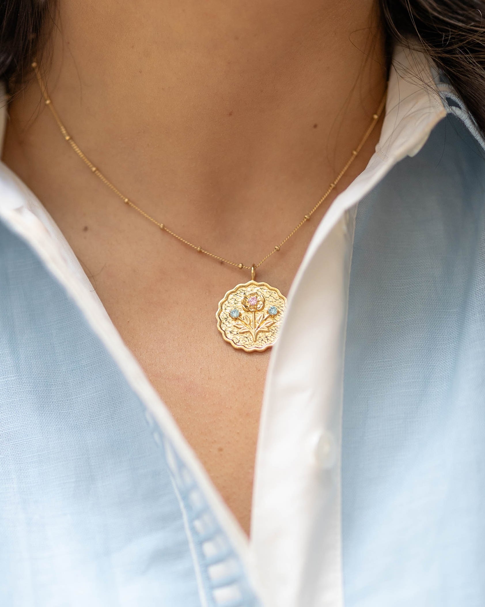 scalloped edge round pendant gold charm with a floral bloom and blue and pink gemstones on a gold chain on a woman's neck