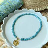 Blue beaded necklace with a gold pendant on a white plate, straw hat, and blue fabric in the background.