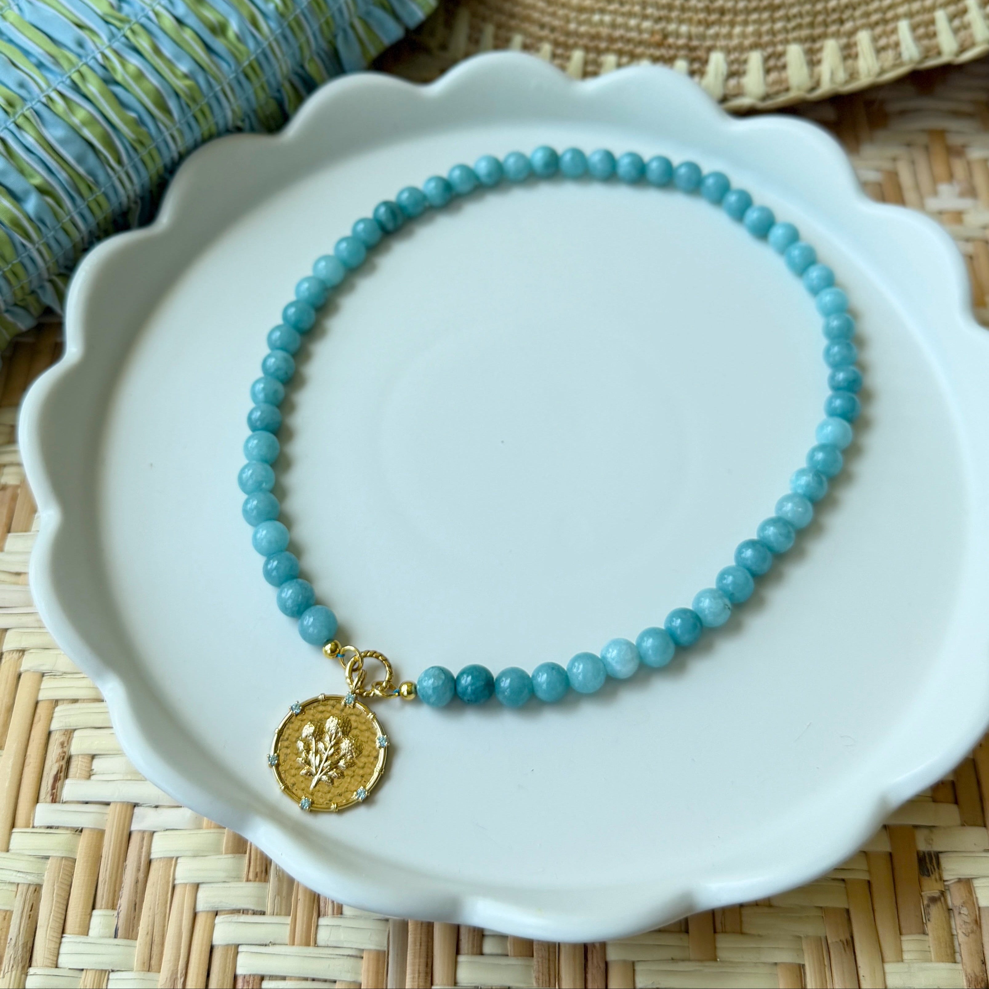 Blue beaded necklace with a gold pendant on a white plate, straw hat, and blue fabric in the background.