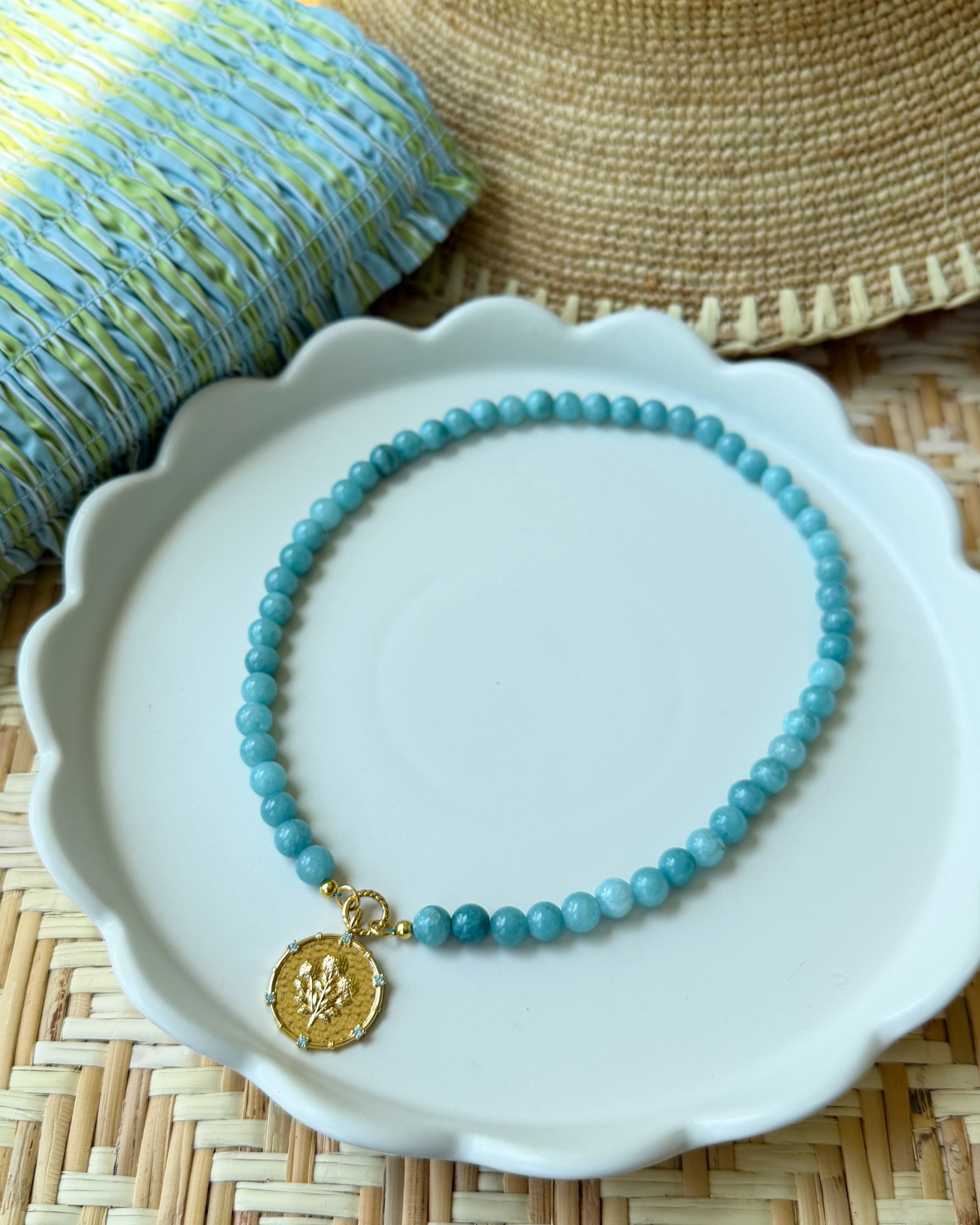 Blue beaded necklace with a gold pendant on a white plate, straw hat, and blue fabric in the background.