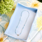 A delicate beaded necklace with light blue beads presented inside an open light blue tray on a floral daisy-patterned fabric surrounded by green hydrangeas.