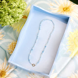 A delicate beaded necklace with light blue beads presented inside an open light blue tray on a floral daisy-patterned fabric surrounded by green hydrangeas.
