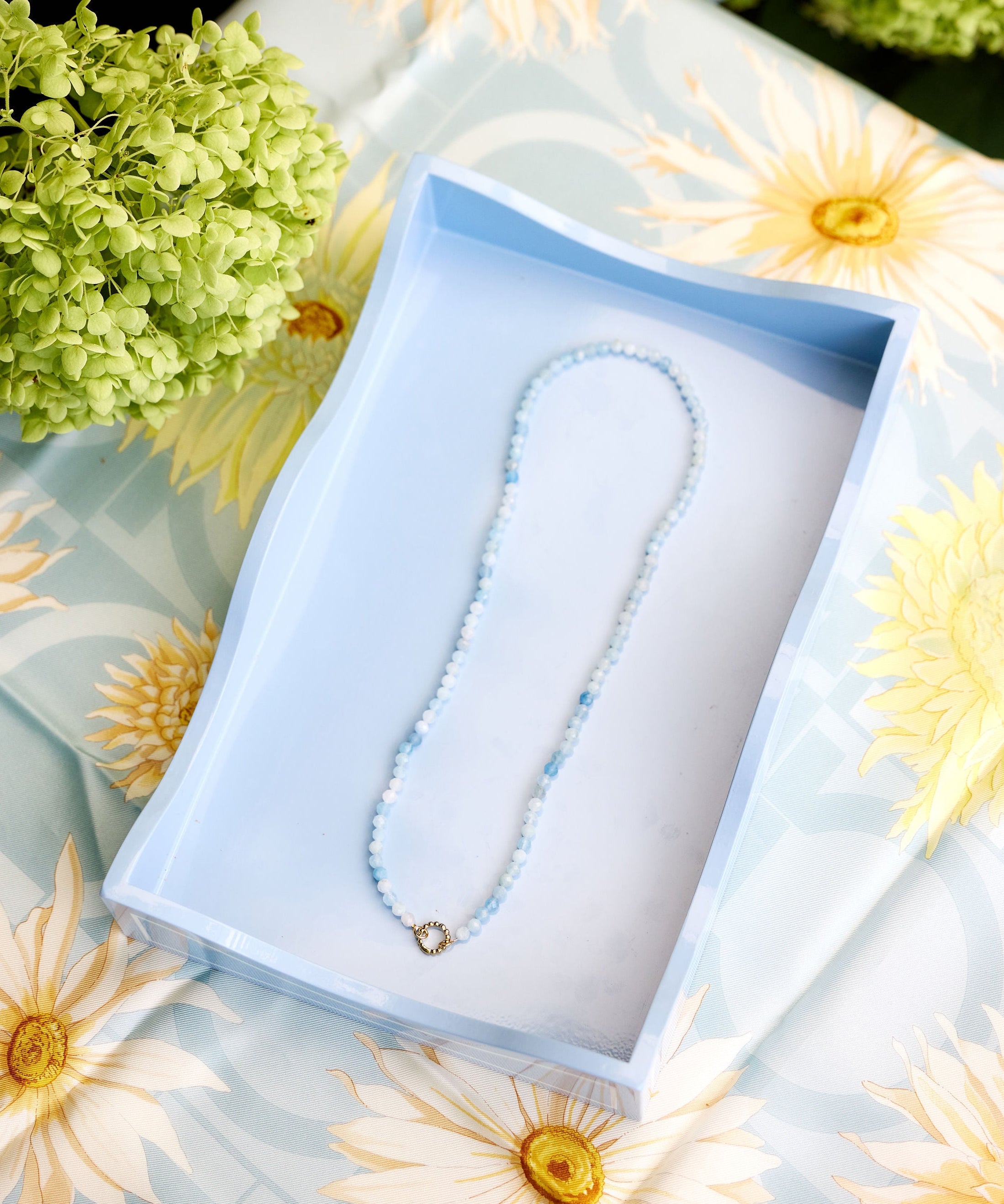 A delicate beaded necklace with light blue beads presented inside an open light blue tray on a floral daisy-patterned fabric surrounded by green hydrangeas.