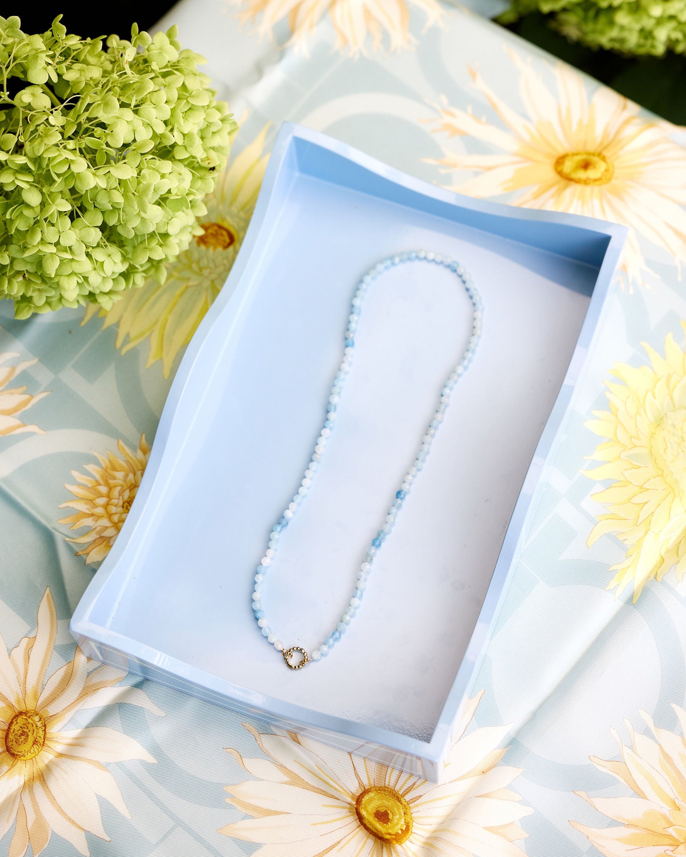 A delicate beaded necklace with light blue beads presented inside an open light blue tray on a floral daisy-patterned fabric surrounded by green hydrangeas.
