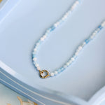 A delicate beaded necklace with light blue beads, featuring a small gold ring clasp, displayed on a light blue tray.