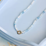 A delicate beaded necklace with light blue beads, featuring a small gold ring clasp, displayed on a light blue tray.