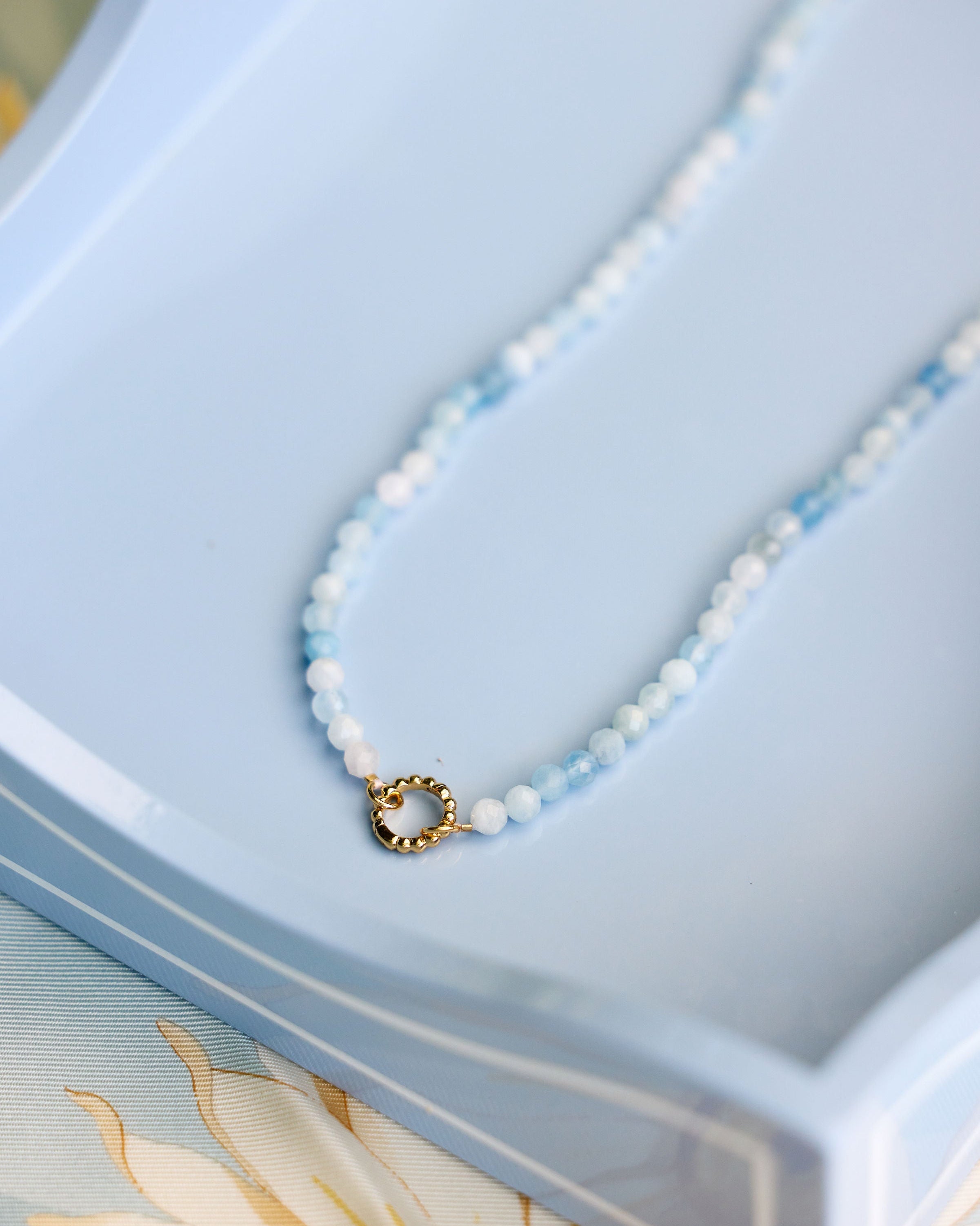 A delicate beaded necklace with light blue beads, featuring a small gold ring clasp, displayed on a light blue tray.