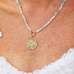 A close-up of a woman wearing a delicate necklace with light blue beads and a round gold pendant, paired with a white lace top.
