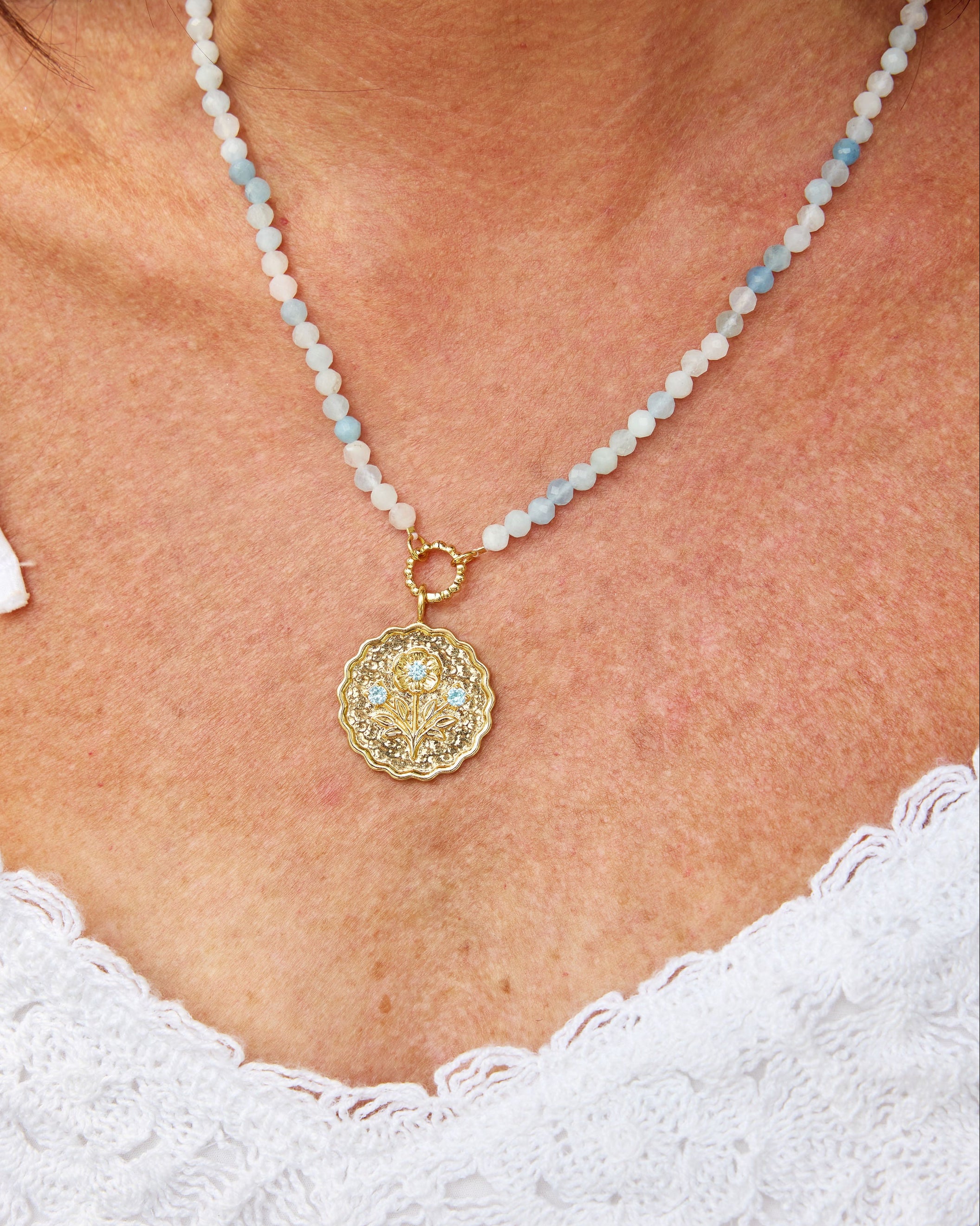 A close-up of a woman wearing a delicate necklace with light blue beads and a round gold pendant, paired with a white lace top.