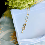 A delicate gold key pendant necklace draped over a light blue ceramic dish, styled with green hydrangea flowers and a light blue patterned fabric background.