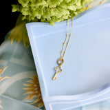 A delicate gold key pendant necklace draped over a light blue ceramic dish, styled with green hydrangea flowers and a light blue patterned fabric background.