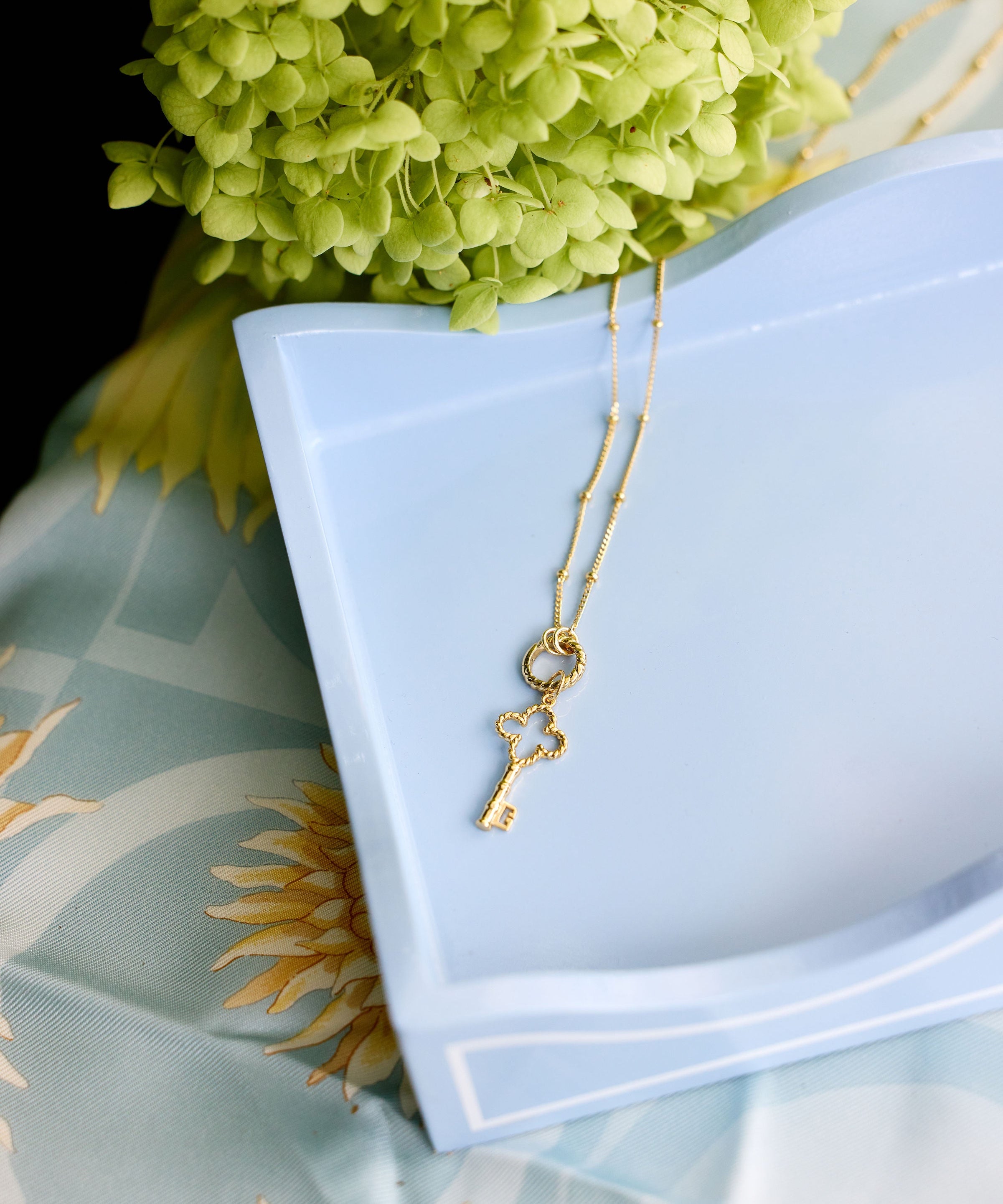 A delicate gold key pendant necklace draped over a light blue ceramic dish, styled with green hydrangea flowers and a light blue patterned fabric background.