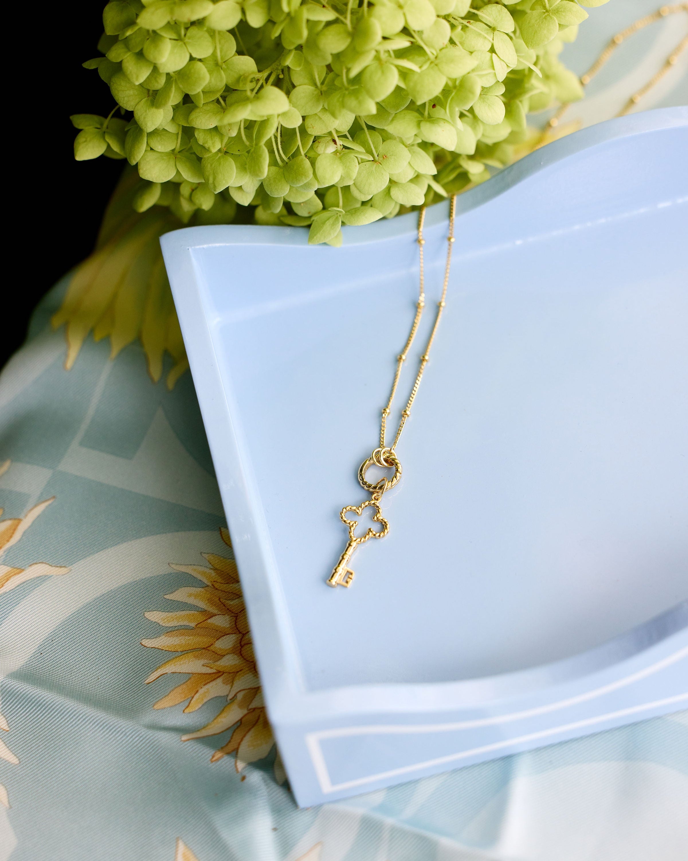 A delicate gold key pendant necklace draped over a light blue ceramic dish, styled with green hydrangea flowers and a light blue patterned fabric background.