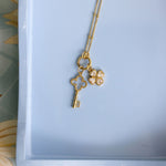 A gold necklace with a key and four-leaf clover pendant, displayed on a light blue tray.
