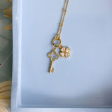 A gold necklace with a key and four-leaf clover pendant, displayed on a light blue tray.