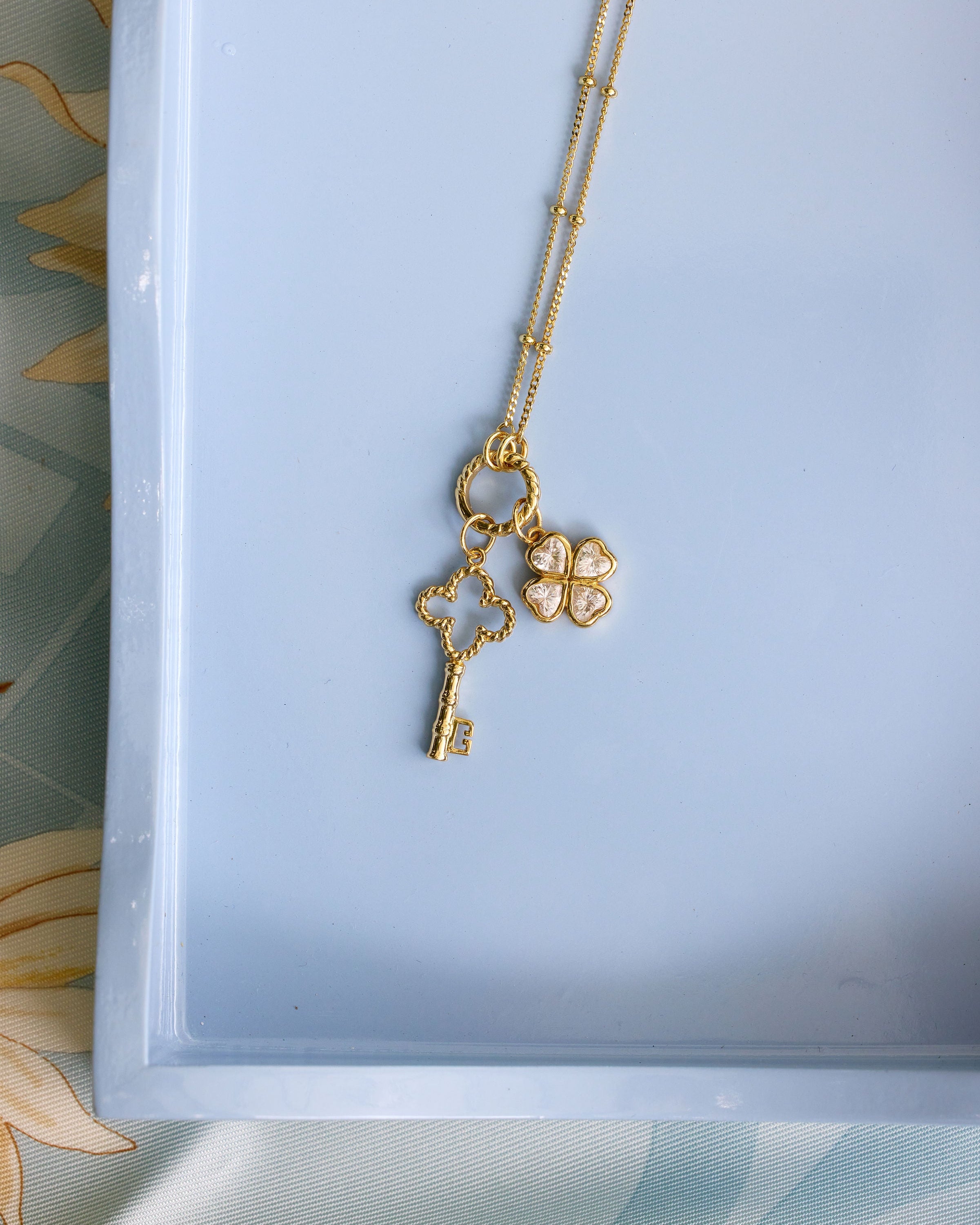 A gold necklace with a key and four-leaf clover pendant, displayed on a light blue tray.