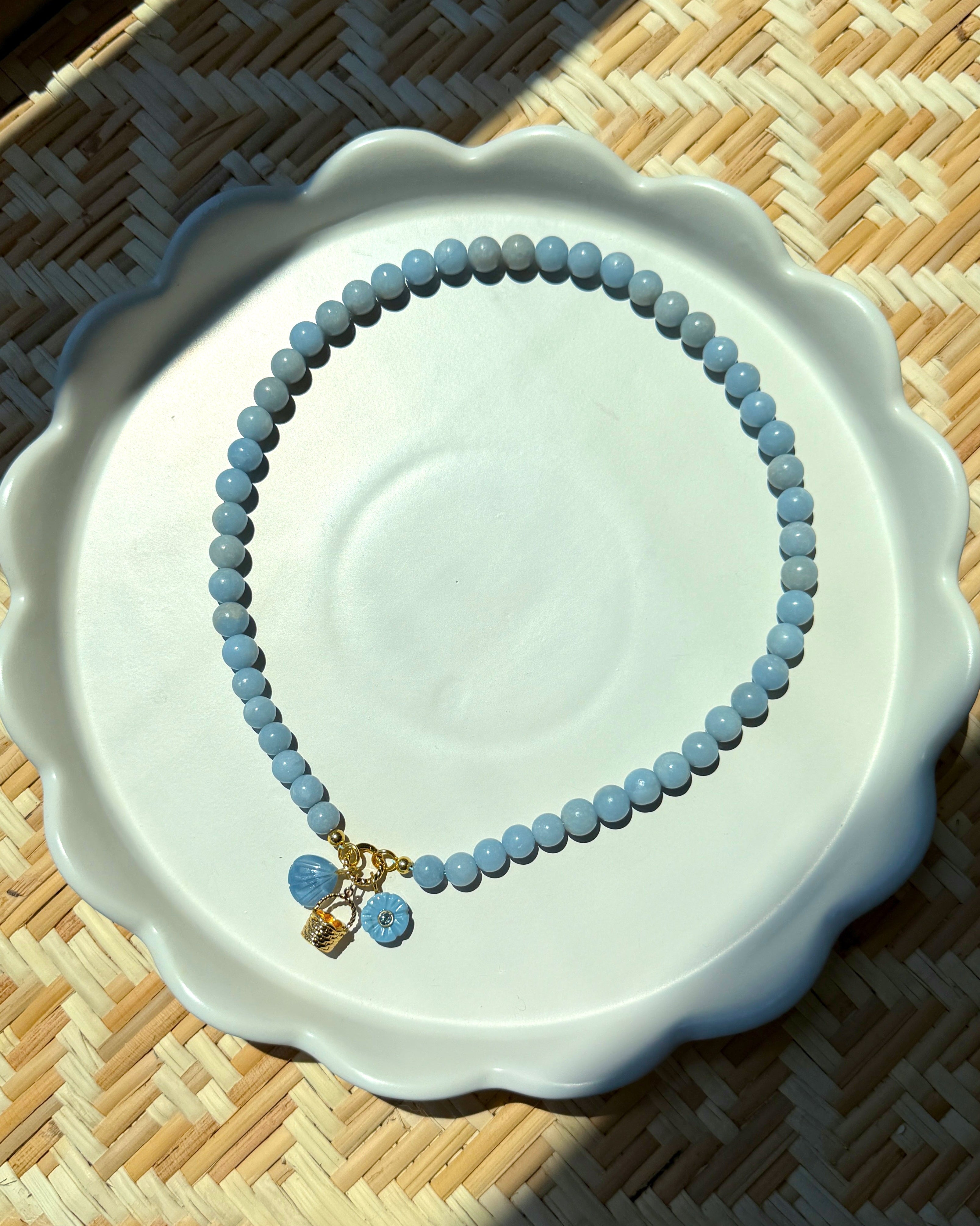 basket charm, blue flower charm, and a blue shell charm on a blue beaded necklace on a white plate