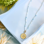 A delicate necklace with a gold-toned medallion pendant and light blue beads, displayed on a light blue tray with floral props.