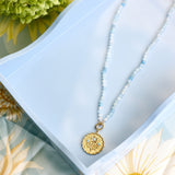 A delicate necklace with a gold-toned medallion pendant and light blue beads, displayed on a light blue tray with floral props.