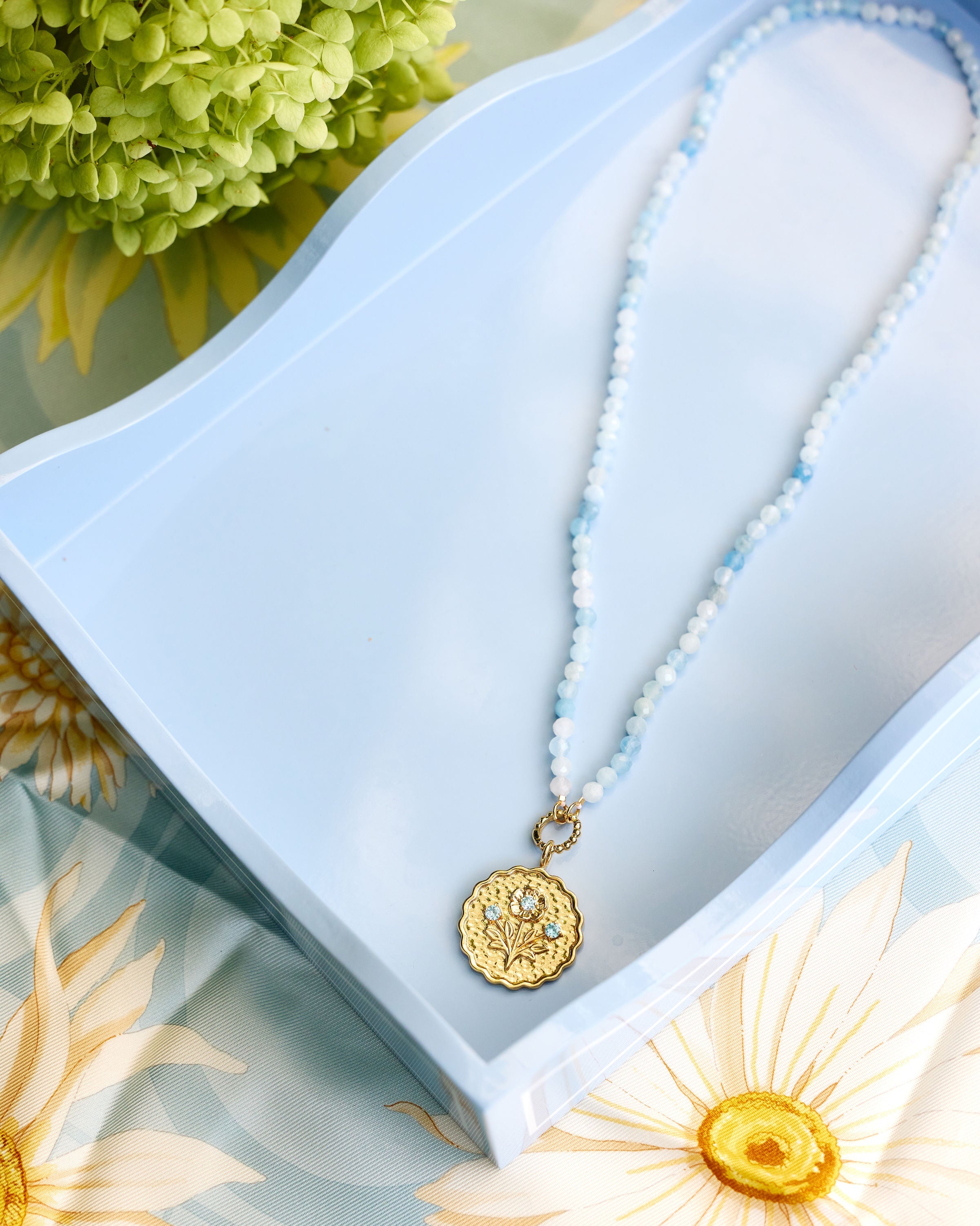 A delicate necklace with a gold-toned medallion pendant and light blue beads, displayed on a light blue tray with floral props.