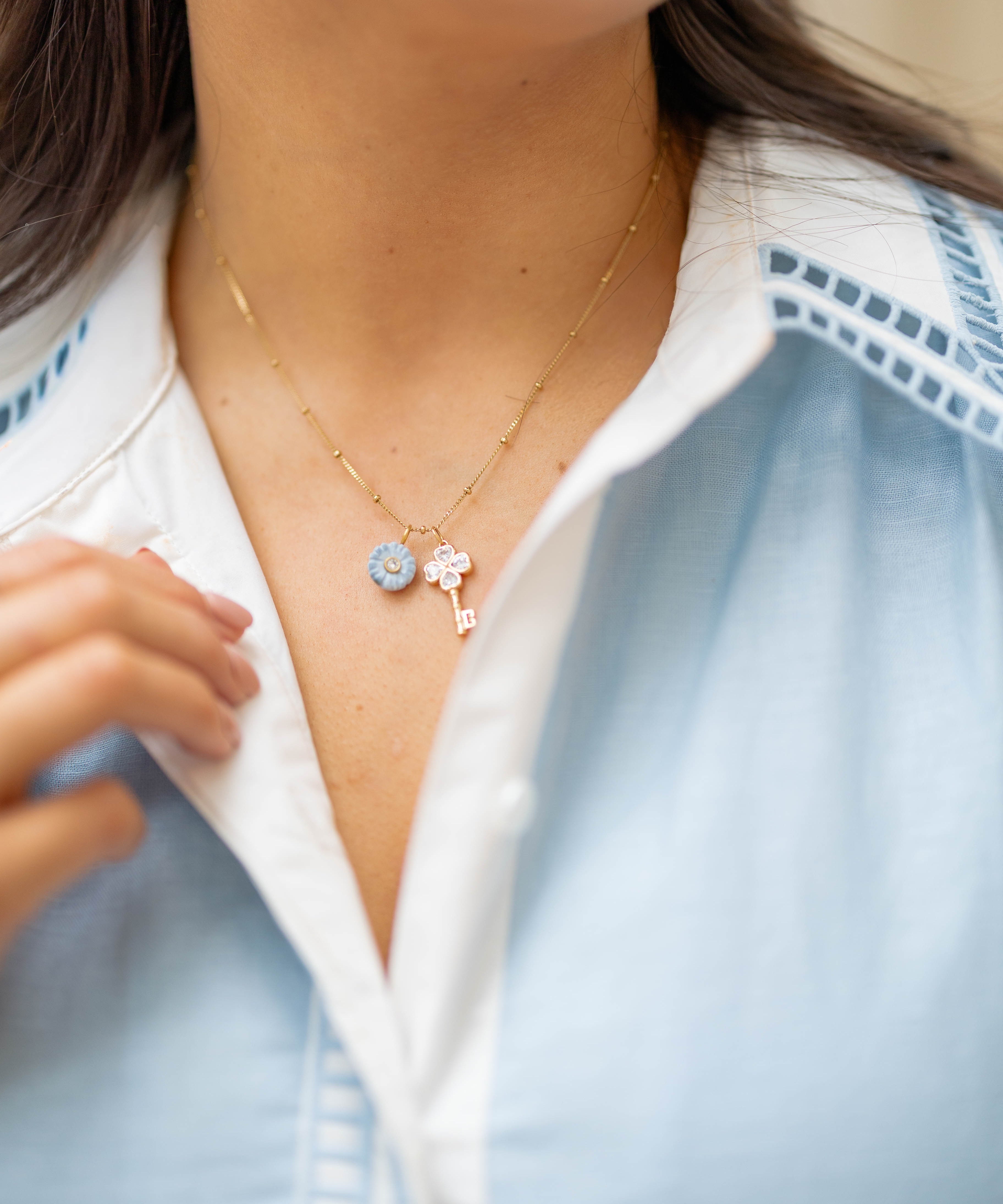 A close-up of a person wearing a delicate gold necklace with a light blue flower charm and key charm