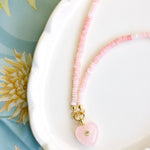 A delicate pink beaded necklace with a pink heart charm resting on a white scalloped dish over a light blue floral fabric background.