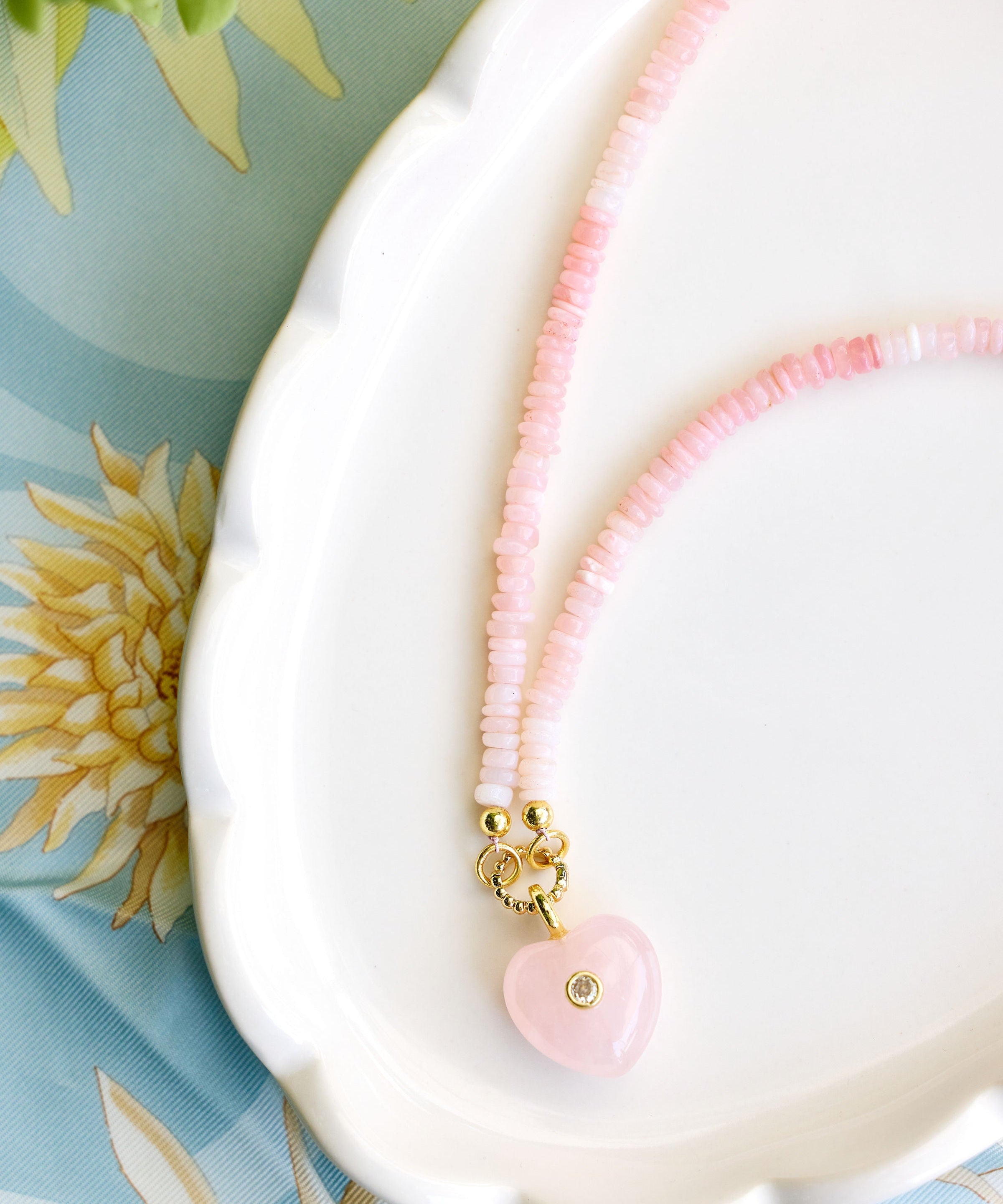 A delicate pink beaded necklace with a pink heart charm resting on a white scalloped dish over a light blue floral fabric background.