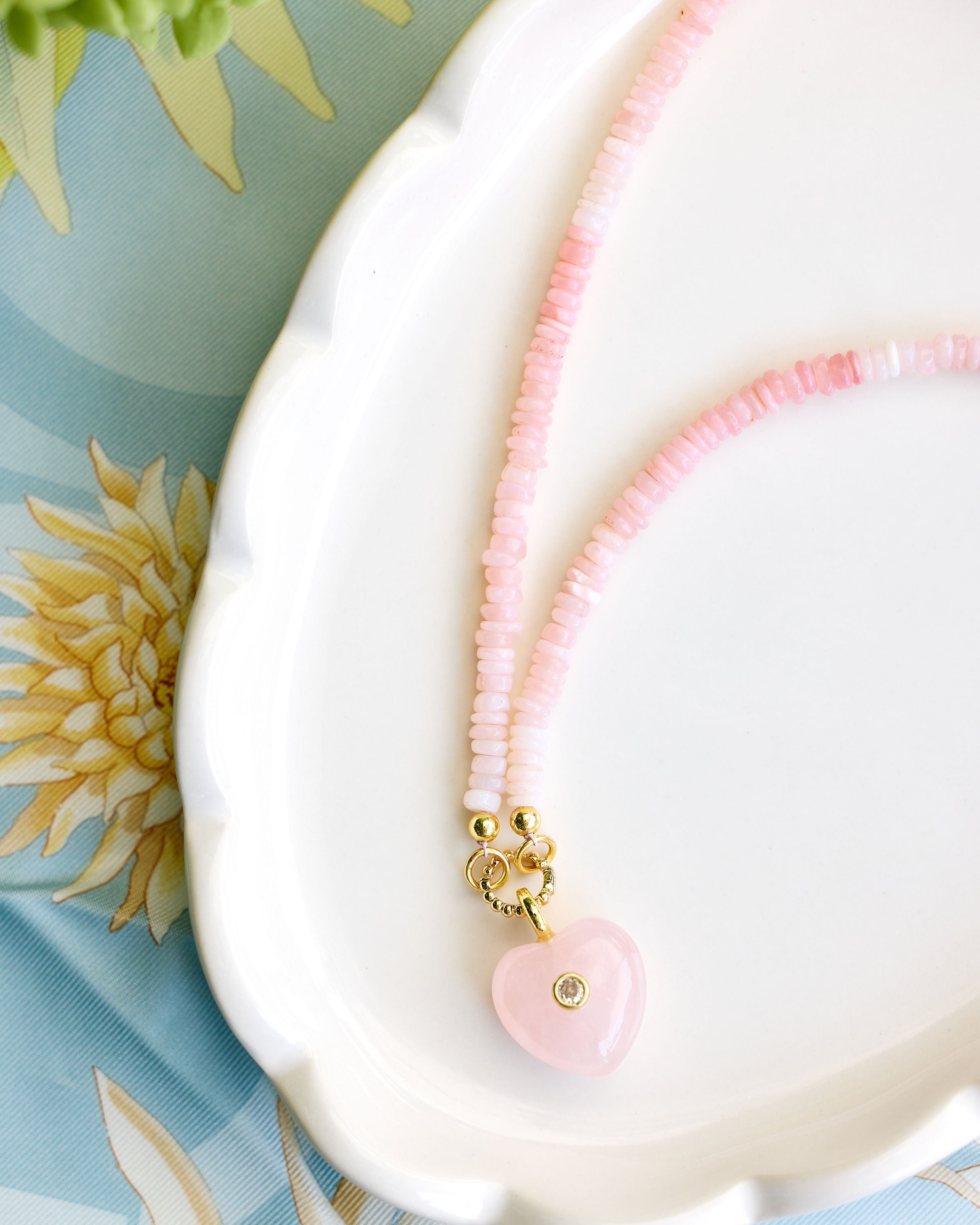A delicate pink beaded necklace with a pink heart charm resting on a white scalloped dish over a light blue floral fabric background.