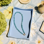 A light blue beaded necklace with a gold clasp rests inside a light blue rectangular box, placed on a pale blue floral tablecloth with decorative plates and greenery around it.
