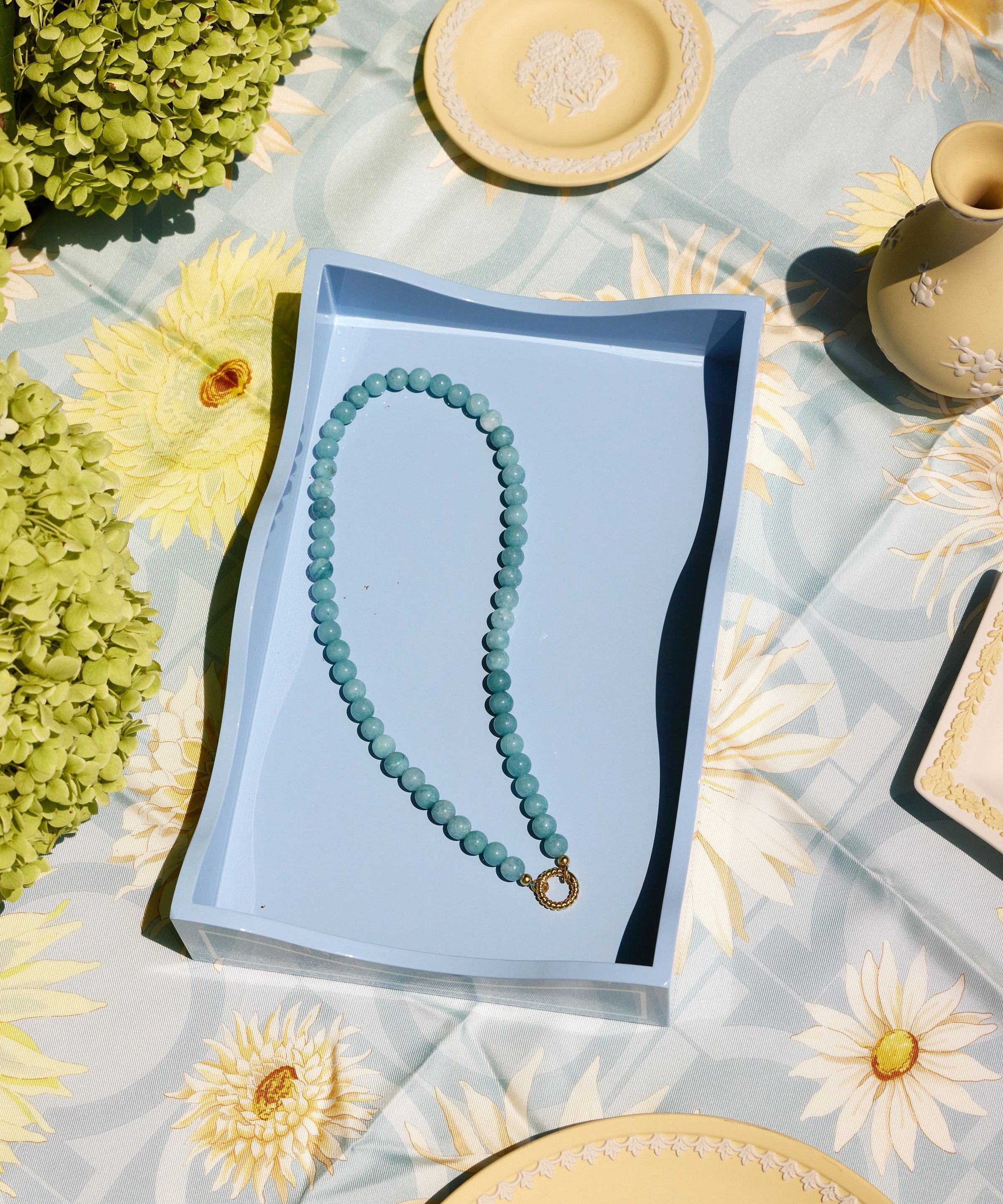 A light blue beaded necklace with a gold clasp rests inside a light blue rectangular box, placed on a pale blue floral tablecloth with decorative plates and greenery around it.