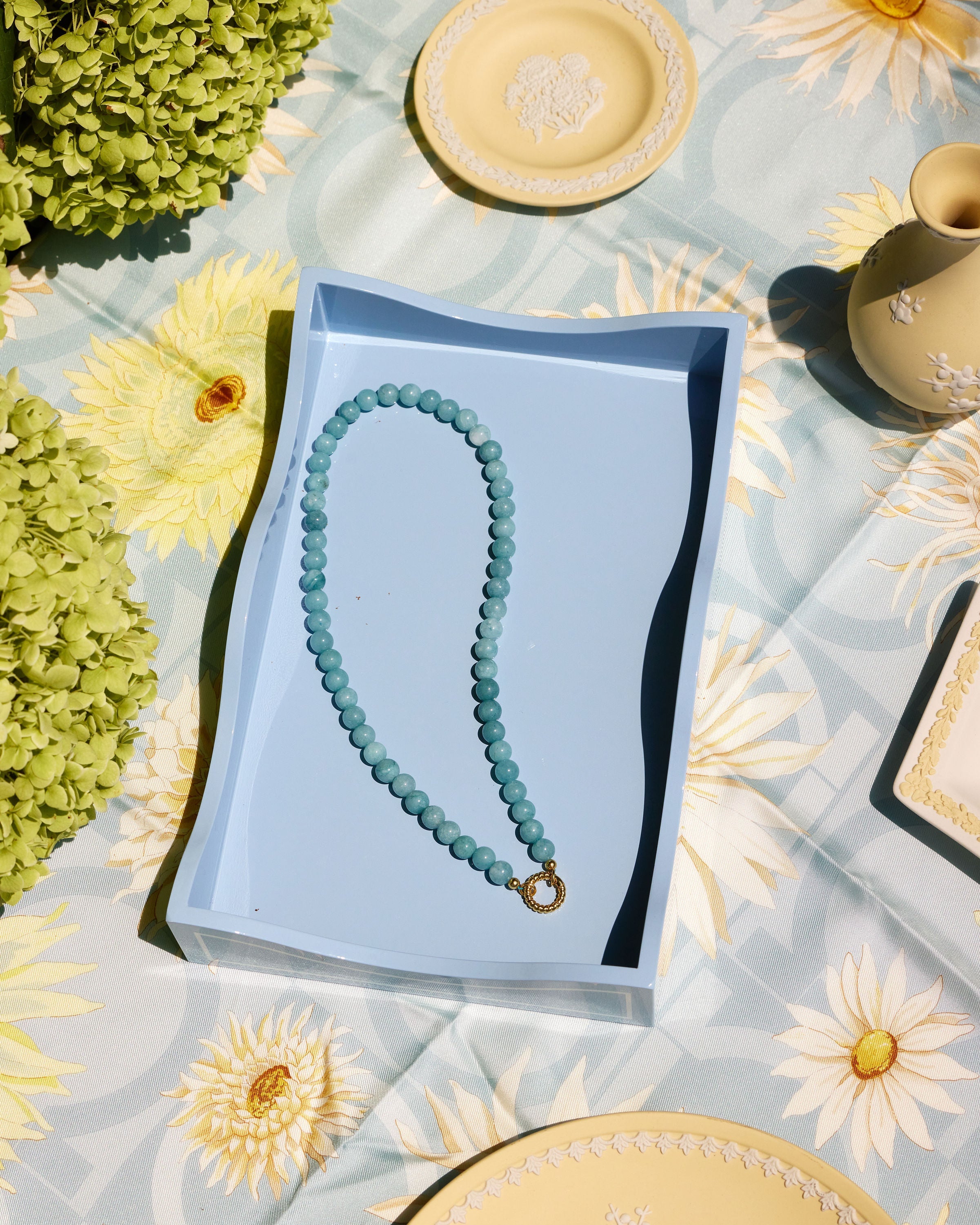 A light blue beaded necklace with a gold clasp rests inside a light blue rectangular box, placed on a pale blue floral tablecloth with decorative plates and greenery around it.