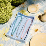 Three beaded necklaces in pastel blue, aqua, and pink are displayed in a light blue box on a floral tablecloth with decorative plates and greenery in the background.