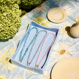 Three beaded necklaces in pastel blue, aqua, and pink are displayed in a light blue box on a floral tablecloth with decorative plates and greenery in the background.