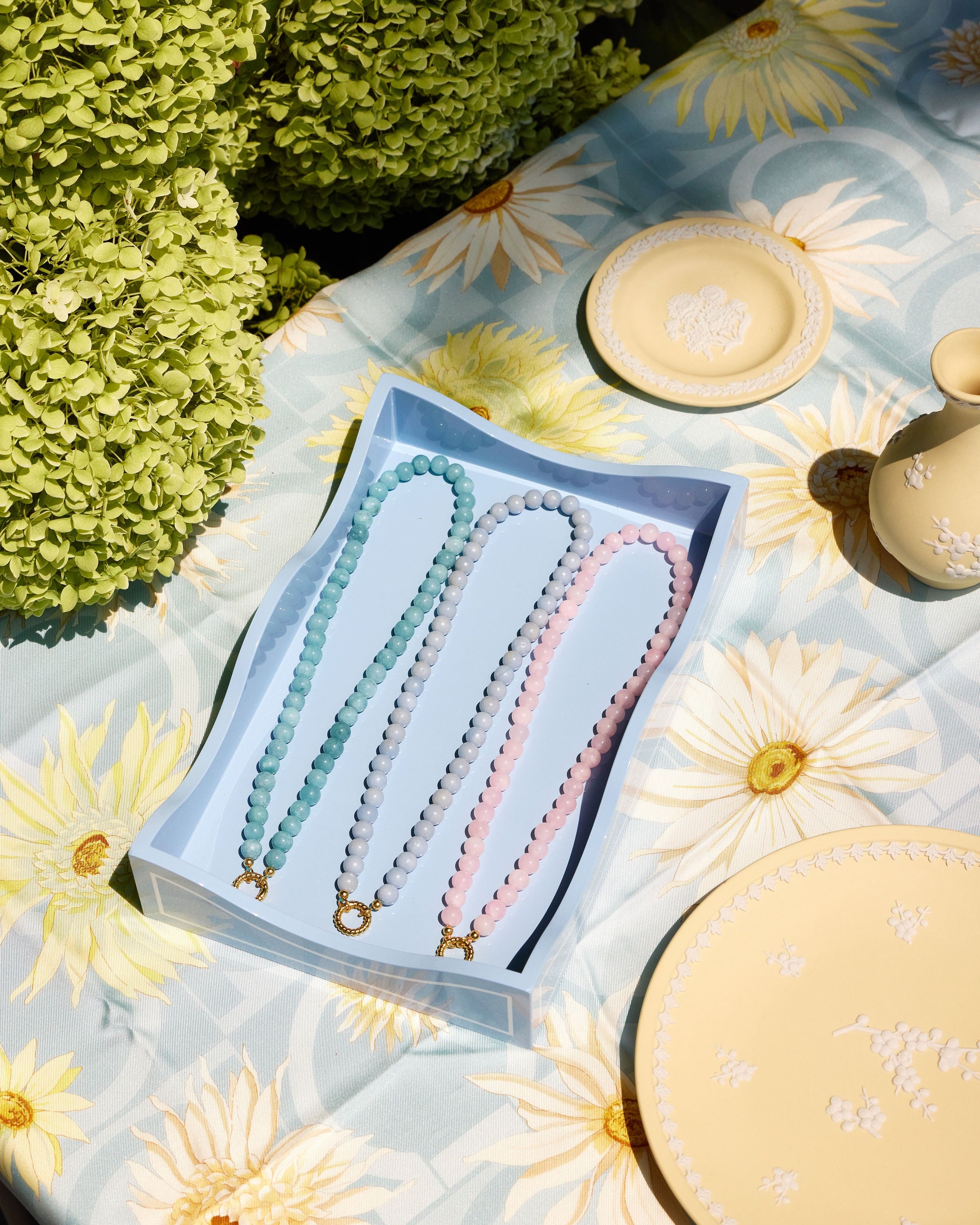 Three beaded necklaces in pastel blue, aqua, and pink are displayed in a light blue box on a floral tablecloth with decorative plates and greenery in the background.