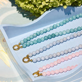 Three beaded jewelry bracelets with gold toggle clasp are displayed on a light blue tray. The beads are in pastel aqua, pale blue, and light pink, with green hydrangea flowers in the background.