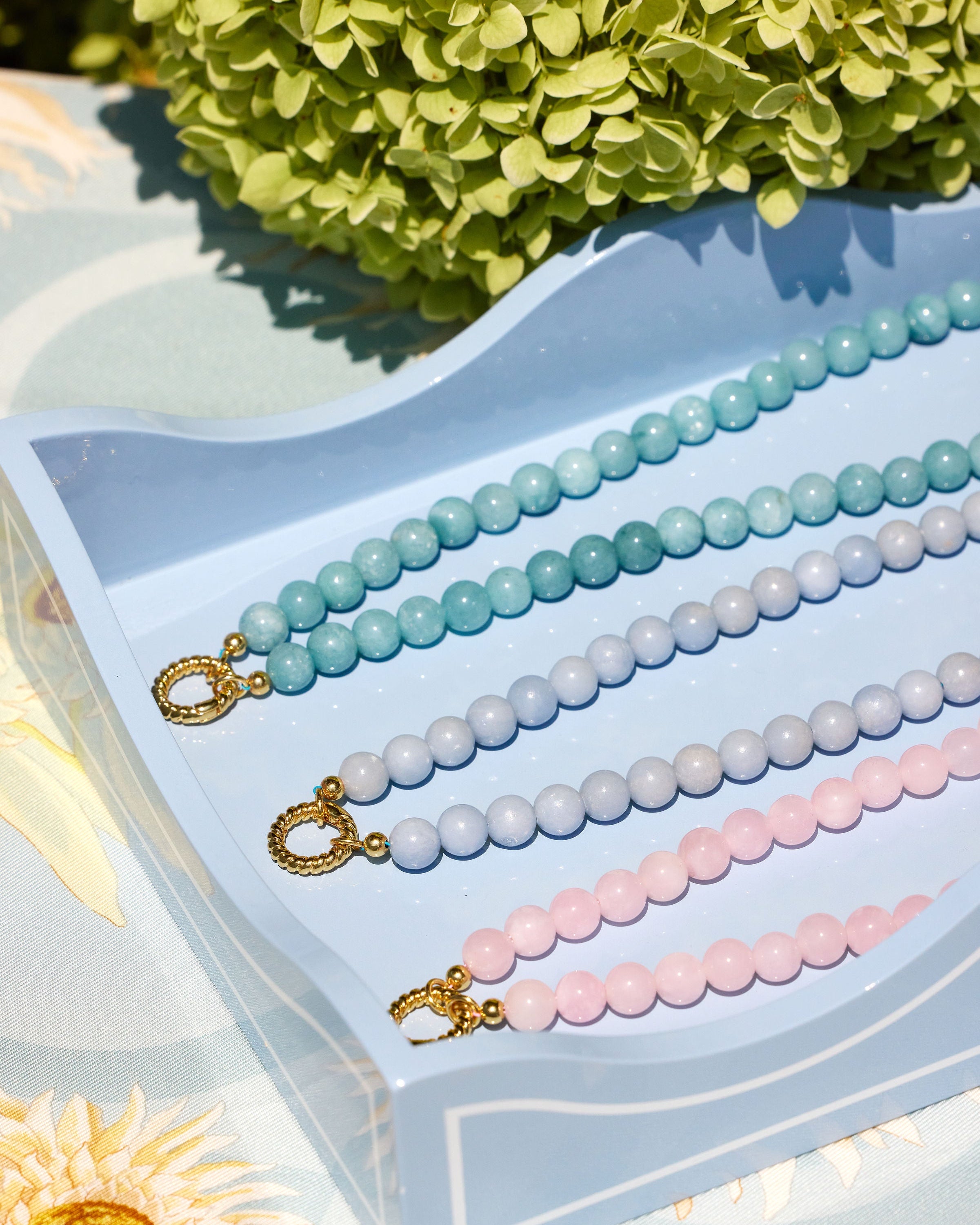 Three beaded jewelry bracelets with gold toggle clasp are displayed on a light blue tray. The beads are in pastel aqua, pale blue, and light pink, with green hydrangea flowers in the background.