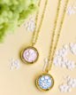 Two gold pendant necklaces with round floral charms, one featuring a pale pink flower and the other a light blue flower, displayed on a cream-colored decorative plate with white floral details and green hydrangeas in the background.