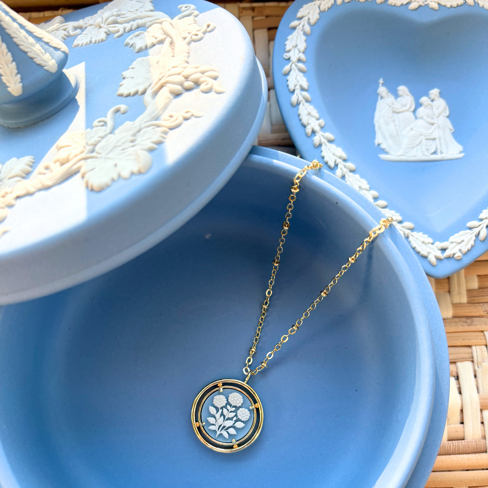 Blue jewelry dish with a gold necklace featuring a circular cameo pendant on a woven surface