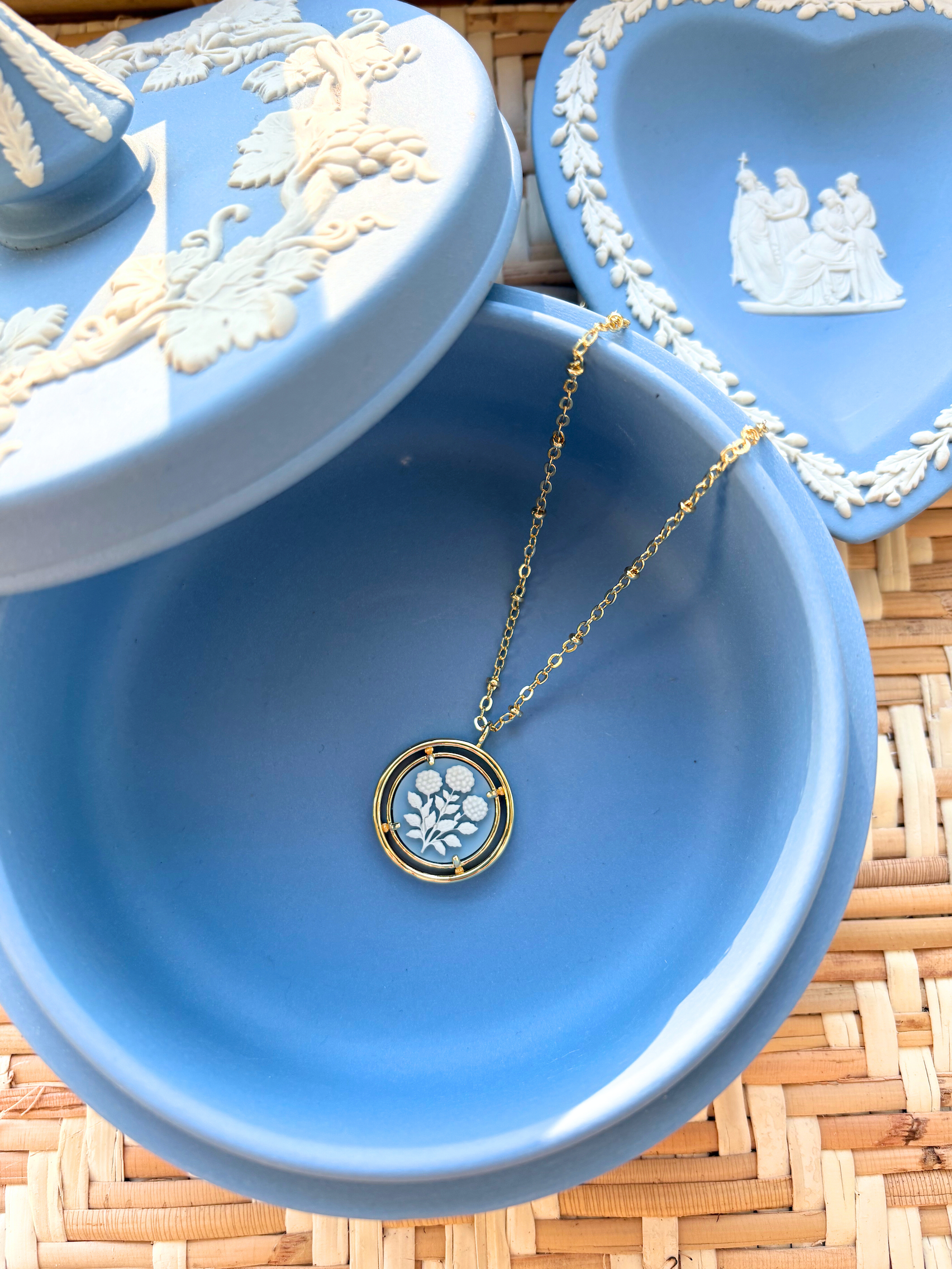 Blue jewelry dish with a gold necklace featuring a circular cameo pendant on a woven surface