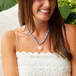 A smiling woman with long brown hair is seated outdoors against a background of green hydrangea flowers. She is wearing a white textured crochet top and a light blue beaded necklace with a white heart-shaped pendant.