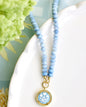 A light blue beaded necklace with a round gold pendant featuring a blue and white floral design, styled on a white ceramic dish with green flowers in the background.